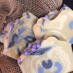 May include: Handmade soap bars with a creamy, marbled design. The soaps have blue and grey swirls, decorated with small purple beads. The bars are displayed on a burlap cloth, highlighting their artisan nature.