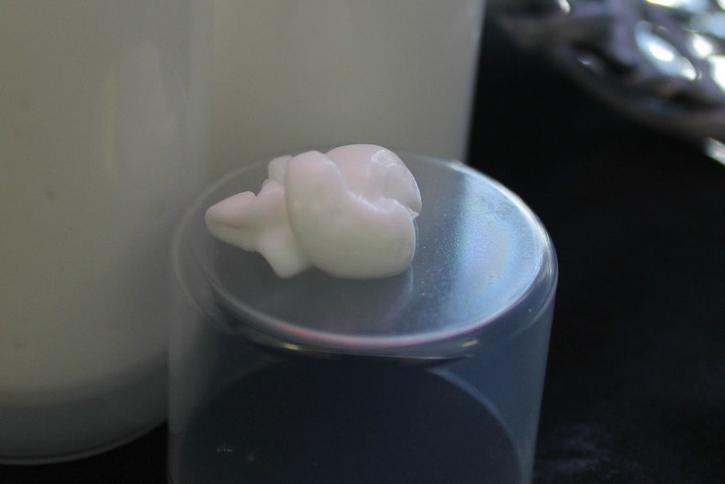 May include: A dollop of thick white cream rests on the translucent, circular cap of a cylindrical container. The cream has a smooth, opaque texture and sits centered on the flat surface of the lid, which is positioned in front of a bottle.