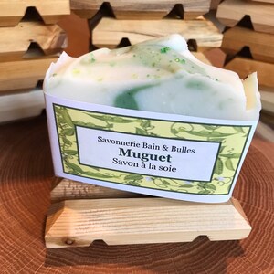 May include: A bar of soap with a white and green swirl design, labeled "Muguet Savon &agrave; la soie." The soap sits on a small wooden soap dish. The background features additional wooden soap dishes.