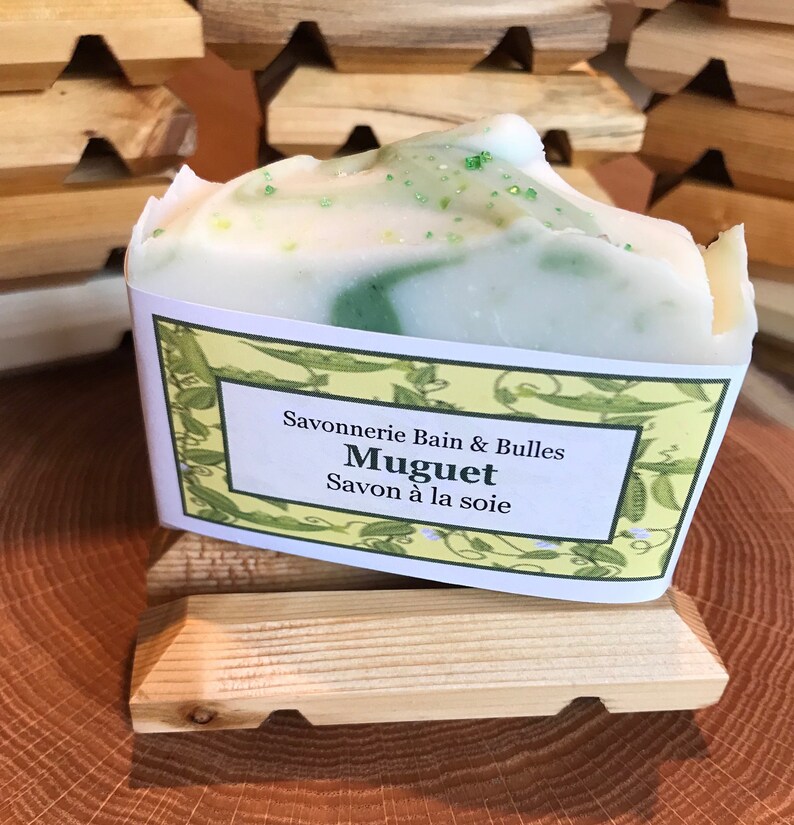 May include: A bar of soap with a white and green swirl design, labeled "Muguet Savon &agrave; la soie." The soap sits on a small wooden soap dish. The background features additional wooden soap dishes.