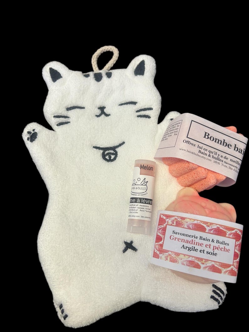 Op de afbeelding: Een witte katvormige washand met zwarte accenten, met een lippenbalsem, een perzikkleurige spons en twee productlabels. De labels luiden "Bombe bai" en "Savonnerie Bain & Bulles Grenadine et p&ecirc;che Argile et soie".