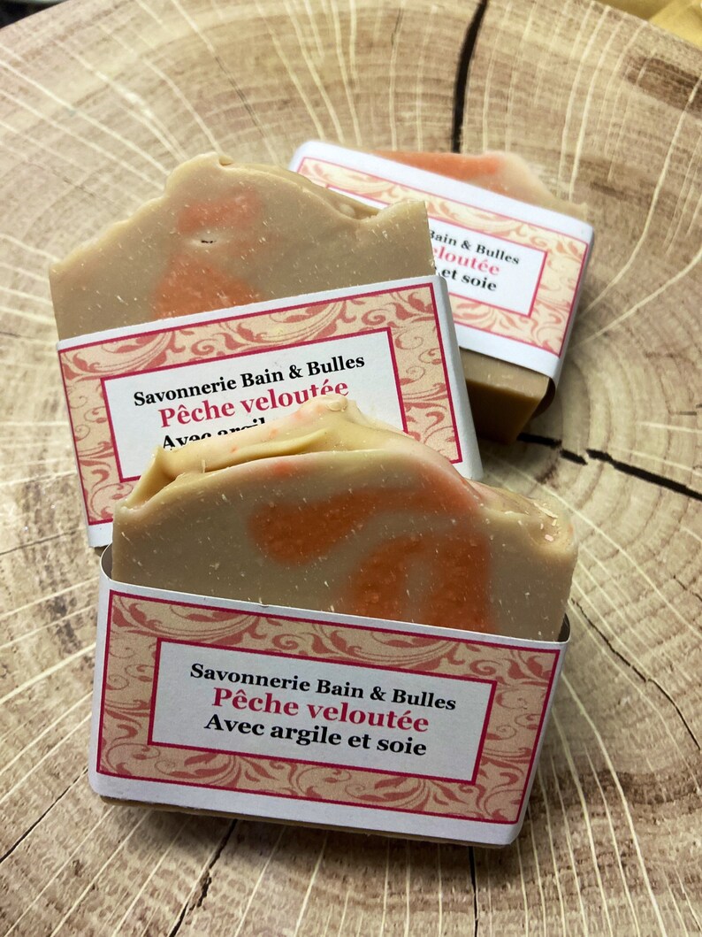 K&ouml;nnte beinhalten: Zwei Seifenst&uuml;cke, die in wei&szlig;es Papier mit rosa und wei&szlig;en Blumenmustern verpackt sind. Die Seife ist mit "Savonnerie Bain & Bulles P&ecirc;che velout&eacute;e Avec argile et soie" beschriftet, was &uuml;bersetzt "Seifenmanufaktur Bain & Bulles Samtpfirsich mit Ton und Seide" bedeutet.