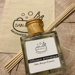 May include: A clear glass bottle with a wooden cap and a white label that reads "Diffuseur aromatique bonheur odorant pour la maison" and "Bain & Bulles Savonnerie Artisanale". The bottle is sitting on a wooden surface with a white cloth bag and wooden sticks.