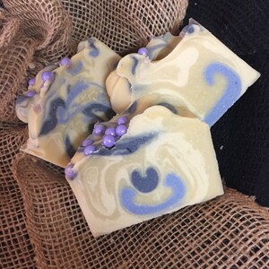 May include: Three handmade soap bars with a creamy white and blue swirl design, accented with small purple beads. The soaps are arranged on a burlap cloth, creating a rustic aesthetic. The soaps have a unique, decorative appearance.