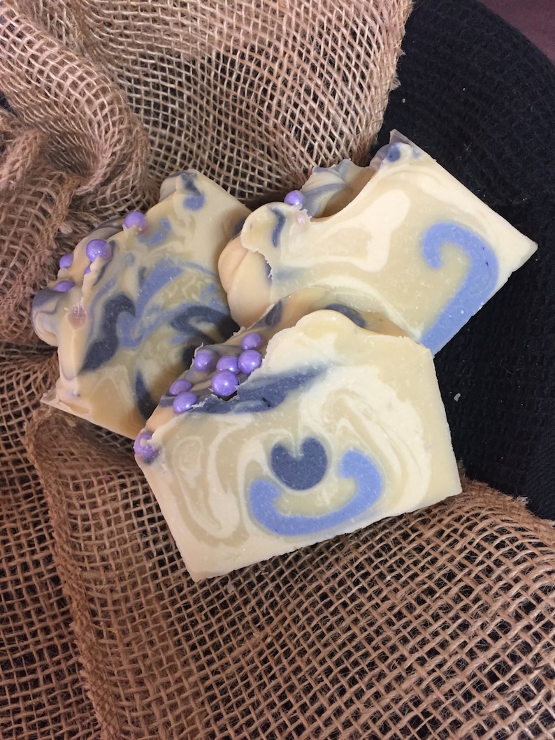 May include: Three handmade soap bars with a creamy white and blue swirl design, accented with small purple beads. The soaps are arranged on a burlap cloth, creating a rustic aesthetic. The soaps have a unique, decorative appearance.