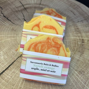 May include: Two bars of handmade soap wrapped in colourful paper. The soap is yellow and orange with swirls. The label reads "Savonnerie Bain & Bulles Neroli orange argile, miel et soie".