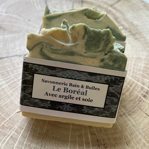 May include: Handmade soap bars with a marbled green and cream design. The soap is wrapped with a label that reads "Savonnerie Bain & Bulles Le Boréal Avec argile et soie."