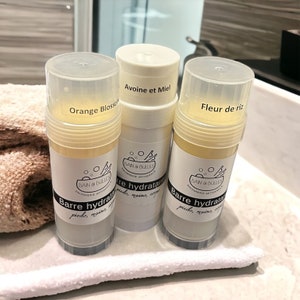 May include: Three white plastic tubes with yellow lotion inside. The tubes are labeled with the text "Orange Blossom", "Avoine et Miel", and "Fleur de riz". Each tube has a black and white label with the text "Bain & Bulles Savonnerie Artisanale" and "Barre hydratante pieds, mains, corps".