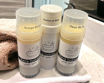 May include: Three white plastic tubes with yellow lotion inside. The tubes are labeled with the text "Orange Blossom", "Avoine et Miel", and "Fleur de riz". Each tube has a black and white label with the text "Bain & Bulles Savonnerie Artisanale" and "Barre hydratante pieds, mains, corps".