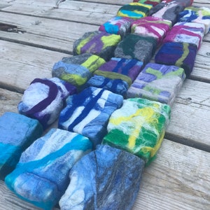 May include: A collection of colorful felted wool soap bars arranged in a row on a wooden surface. The bars are various shades of blue, green, yellow, purple, and pink, with swirls and patterns throughout.