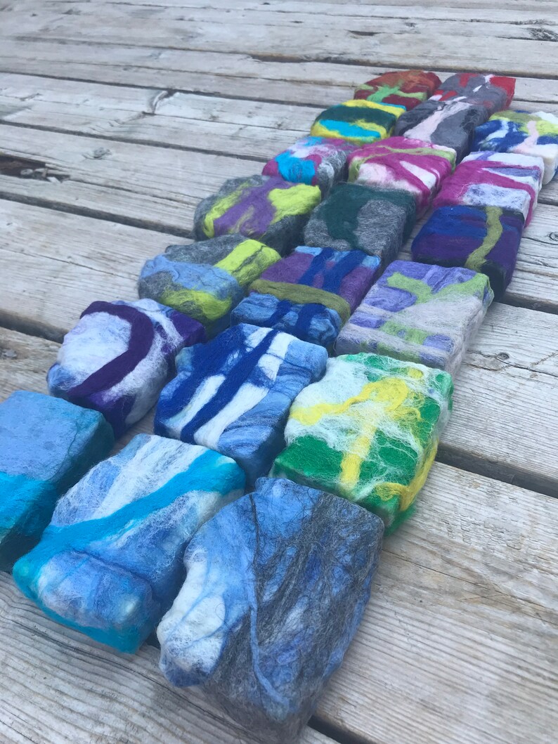May include: A collection of colorful felted wool soap bars arranged in a row on a wooden surface. The bars are various shades of blue, green, yellow, purple, and pink, with swirls and patterns throughout.