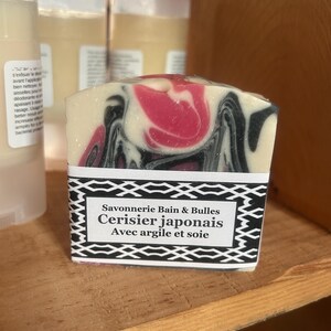Puede incluir: Una pastilla de jab&oacute;n rectangular con un patr&oacute;n jaspeado de remolinos color crema, negro y rosa. Una etiqueta de papel blanco con un borde geom&eacute;trico negro presenta el texto Savonnerie Bain & Bulles, Cerisier japonais y Avec argile et soie.