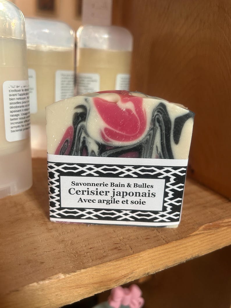 Puede incluir: Una pastilla de jab&oacute;n rectangular con un patr&oacute;n jaspeado de remolinos color crema, negro y rosa. Una etiqueta de papel blanco con un borde geom&eacute;trico negro presenta el texto Savonnerie Bain & Bulles, Cerisier japonais y Avec argile et soie.