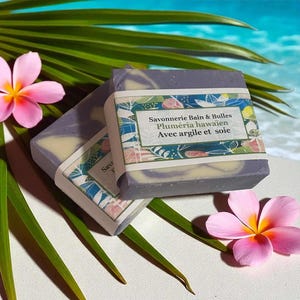 Peut inclure: Deux savons artisanaux avec un tourbillon lavande et crème, enveloppés dans du papier avec des motifs floraux. Le savon est étiqueté "Savonnerie Bain & Bulles Plumeria hawaïen Avec argile et soie". Des fleurs de plumeria roses et des feuilles vertes sont en arrière-plan.