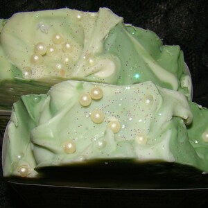 May include: Two bars of handmade soap with a layered design of light and dark green, decorated with small, round, pearl-like embellishments and sparkling glitter. The soap has a swirled, textured appearance.