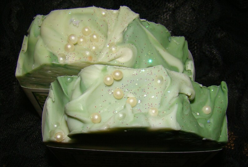 May include: Two bars of handmade soap with a layered design of light and dark green, decorated with small, round, pearl-like embellishments and sparkling glitter. The soap has a swirled, textured appearance.