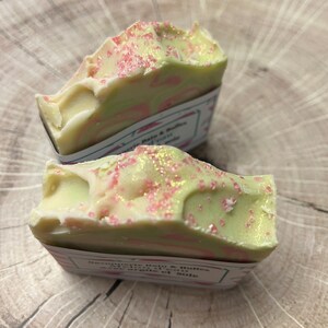 Op de afbeelding: Twee handgemaakte zeepjes met een groen-wit swirl-ontwerp. De zeep is bedekt met roze en gouden glitter. De zeep is gelabeld "Savonnette Palo & Tulasi" en "Aroma de vainilla et soie".