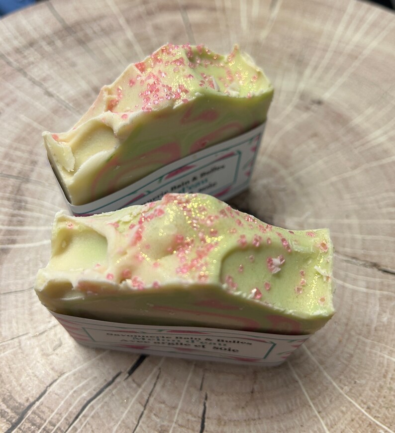 Op de afbeelding: Twee handgemaakte zeepjes met een groen-wit swirl-ontwerp. De zeep is bedekt met roze en gouden glitter. De zeep is gelabeld "Savonnette Palo & Tulasi" en "Aroma de vainilla et soie".