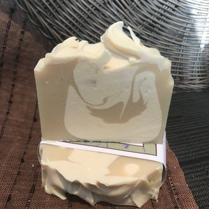 May include: Two bars of handmade soap, one on top of the other. The top bar is a light beige colour with swirls of white. The bottom bar is a similar colour with a white swirl in the centre. Both bars have a slightly textured surface.