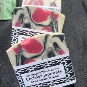 Puede incluir: Tres pastillas de jab&oacute;n rectangulares con un patr&oacute;n jaspeado en crema, negro y rosa brillante. Cada pastilla est&aacute; envuelta en una banda de papel blanco con un borde geom&eacute;trico negro y el texto Savonnerie Bain & Bulles Cerisier japonais Avec argile et soie.