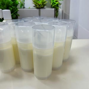 May include: A collection of cylindrical plastic pump bottles filled with an opaque off-white cream. Each container features a clear, removable cap and a white pump mechanism, arranged in a cluster on a smooth, light-colored surface.