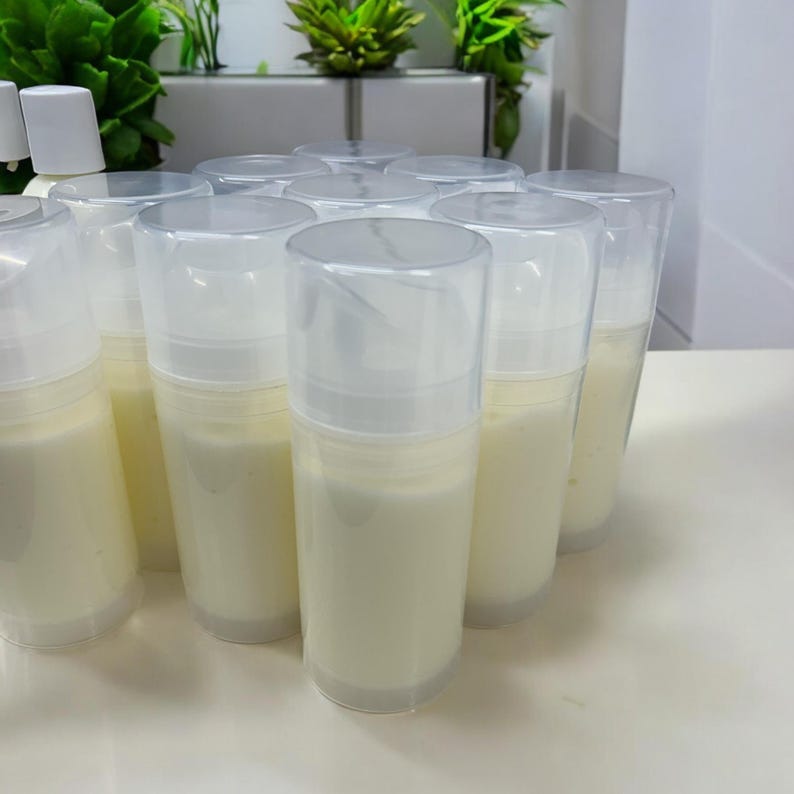 May include: A collection of cylindrical plastic pump bottles filled with an opaque off-white cream. Each container features a clear, removable cap and a white pump mechanism, arranged in a cluster on a smooth, light-colored surface.