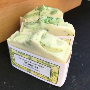 May include: Three bars of handmade soap with a creamy white and light green swirl design. The soap is labeled "Muguet Savon &agrave; la soie" and has a light green sprinkle on top. The soap is wrapped in a white label with a yellow floral design.