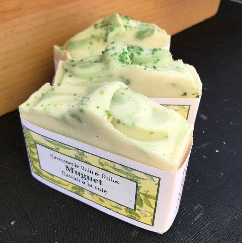 May include: Three bars of handmade soap with a creamy white and light green swirl design. The soap is labeled "Muguet Savon &agrave; la soie" and has a light green sprinkle on top. The soap is wrapped in a white label with a yellow floral design.