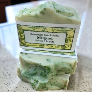 May include: Two bars of handmade soap with a light green and white color scheme. The soap has a textured surface and is speckled with green particles. Each bar is wrapped with a label that reads "Savonnerie Bain & Bulles Muguet Savon &agrave; la soie."