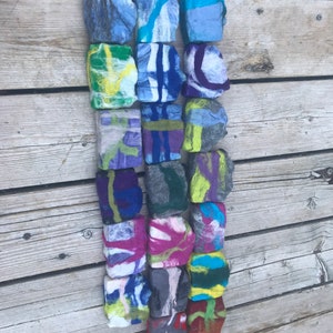 May include: A collection of 18 felted soap bars in various colors and patterns, including blue, green, purple, red, and yellow. The bars are arranged in a vertical row on a wooden surface.