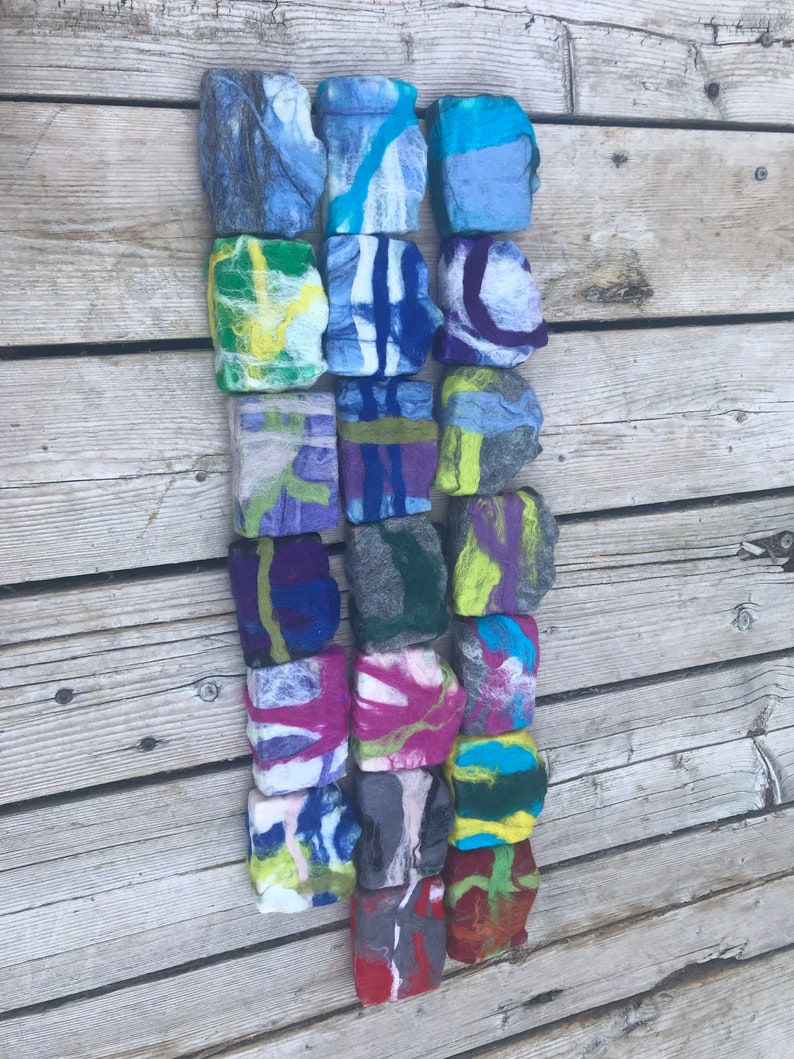 May include: A collection of 18 felted soap bars in various colors and patterns, including blue, green, purple, red, and yellow. The bars are arranged in a vertical row on a wooden surface.