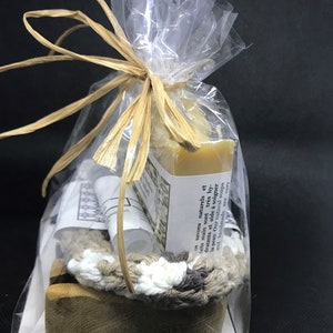 Puede incluir: Una bolsa de pl&aacute;stico transparente llena de una barra de jab&oacute;n natural, una peque&ntilde;a botella de loci&oacute;n, un portajab&oacute;n de ganchillo y un jabonera de madera. La bolsa est&aacute; atada con una cinta de rafia marr&oacute;n. La barra de jab&oacute;n est&aacute; etiquetada con el texto "os savons naturels et faits main sont tr&egrave;s hydratants et aide &agrave; soigner la peau. Our natural soaps and handmade are very"