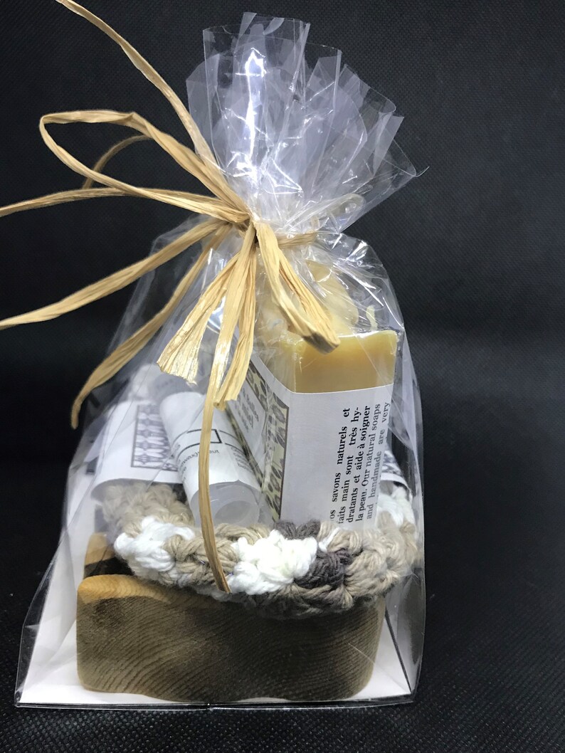 Puede incluir: Una bolsa de pl&aacute;stico transparente llena de una barra de jab&oacute;n natural, una peque&ntilde;a botella de loci&oacute;n, un portajab&oacute;n de ganchillo y un jabonera de madera. La bolsa est&aacute; atada con una cinta de rafia marr&oacute;n. La barra de jab&oacute;n est&aacute; etiquetada con el texto "os savons naturels et faits main sont tr&egrave;s hydratants et aide &agrave; soigner la peau. Our natural soaps and handmade are very"