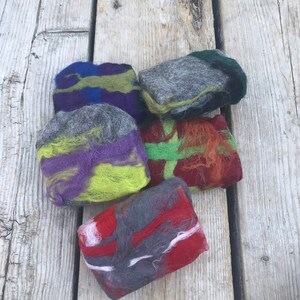 May include: Five handmade soap bars with different color combinations of gray, green, purple, red, and white. The soap bars are made with wool and are arranged on a wooden surface.
