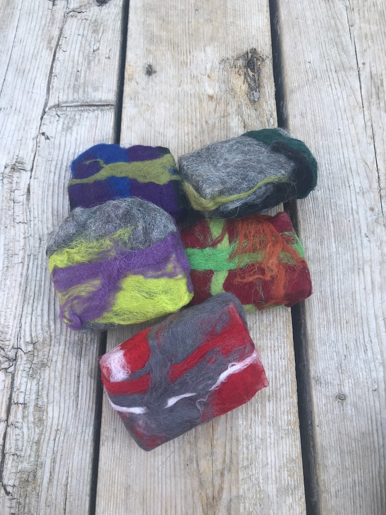 May include: Five handmade soap bars with different color combinations of gray, green, purple, red, and white. The soap bars are made with wool and are arranged on a wooden surface.