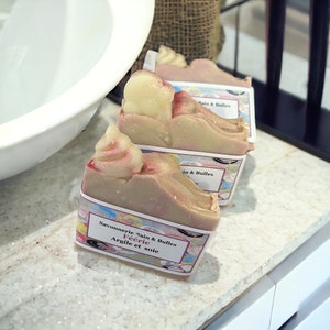 May include: Two bars of handmade soap with a swirl design. The soap is a light brown color with pink swirls. The soap is labeled "Savonnerie Bain & Bulles Féeerie Argile et Soie".