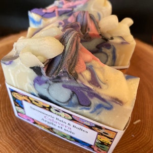 May include: Two bars of handmade soap with a swirl design in shades of purple, blue, and white. The soap is labelled "Savonnerie Bain & Bulles Calavera Argile et soie".