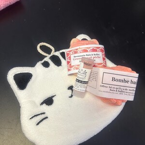 Op de afbeelding: Een witte katvormige washand met zwarte geborduurde details, met een lippenbalsem tube en twee roze badproducten met labels. De labels hebben Franse tekst. De washand heeft een lus om op te hangen.