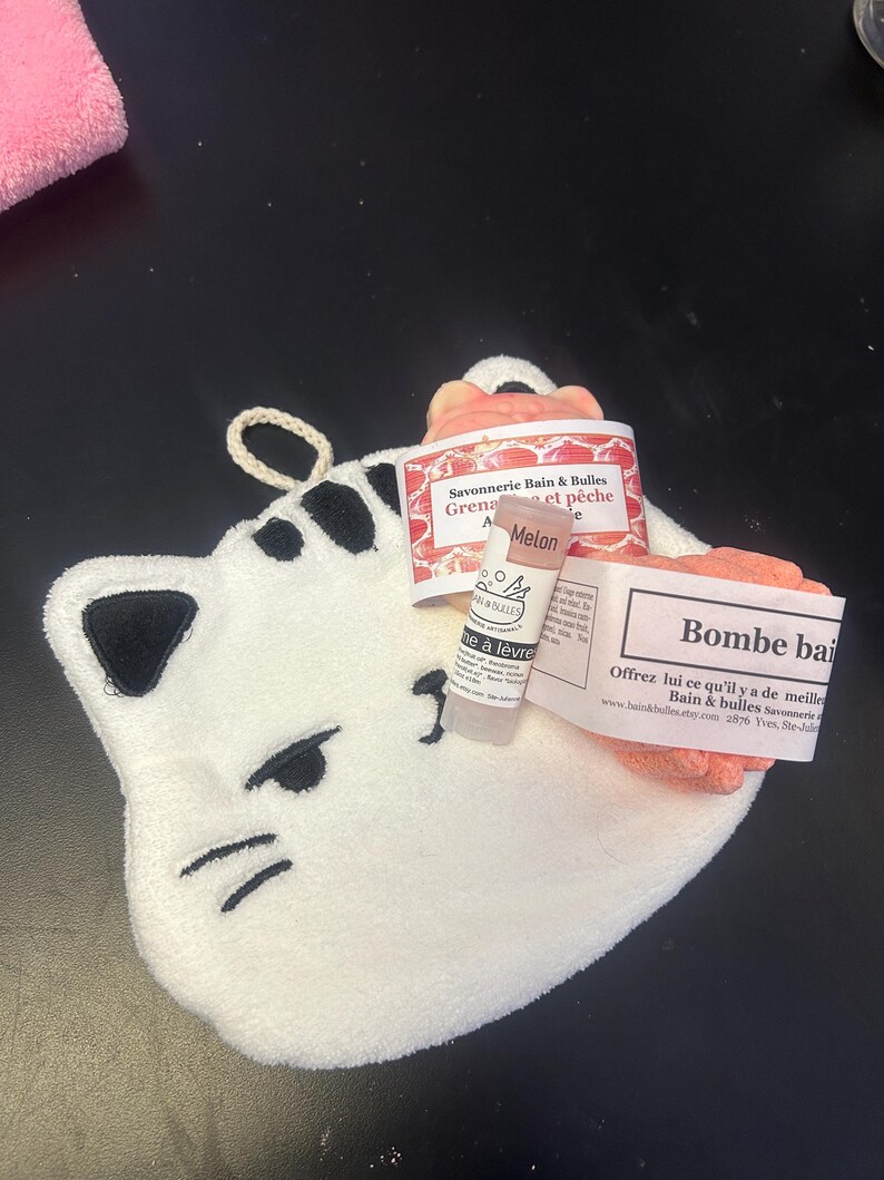 Op de afbeelding: Een witte katvormige washand met zwarte geborduurde details, met een lippenbalsem tube en twee roze badproducten met labels. De labels hebben Franse tekst. De washand heeft een lus om op te hangen.