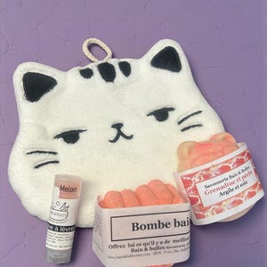 Op de afbeelding: Een witte katvormige washand met zwarte accenten, een lippenbalsem en twee dozen met badproducten. De lippenbalsem is voorzien van het label "Melon". De badproducten zijn voorzien van de labels "Bombe bai" en "Savonnerie Bain & Bulles Grenadine et p&ecirc;che Argile et soie".