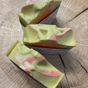 May include: Three bars of handmade soap with a light green base, swirled with pink and orange hues. The soap has a pearlescent sheen and is displayed on a wooden surface. The soap appears to be freshly cut.