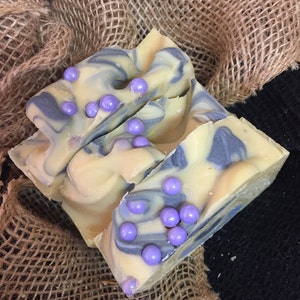 May include: Handmade soap bars with a creamy yellow base, swirled with blue-grey hues. The soap is decorated with small, round, lavender-coloured beads. The soap rests on a burlap surface, creating a rustic aesthetic.
