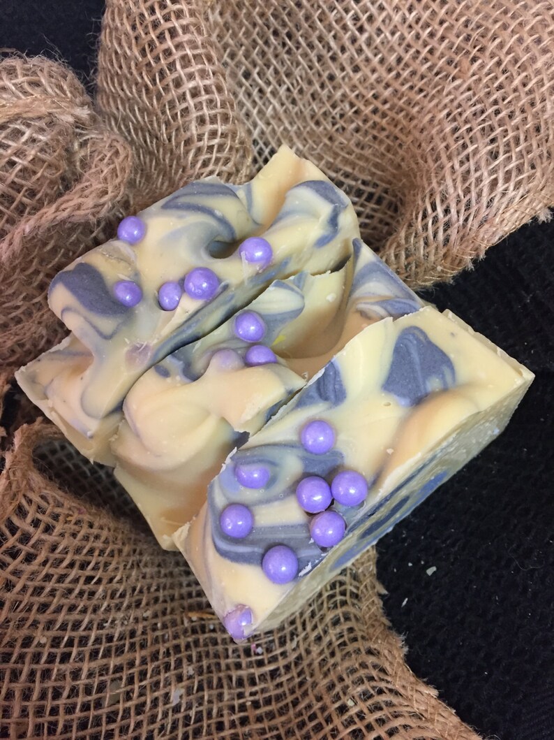 May include: Handmade soap bars with a creamy yellow base, swirled with blue-grey hues. The soap is decorated with small, round, lavender-coloured beads. The soap rests on a burlap surface, creating a rustic aesthetic.