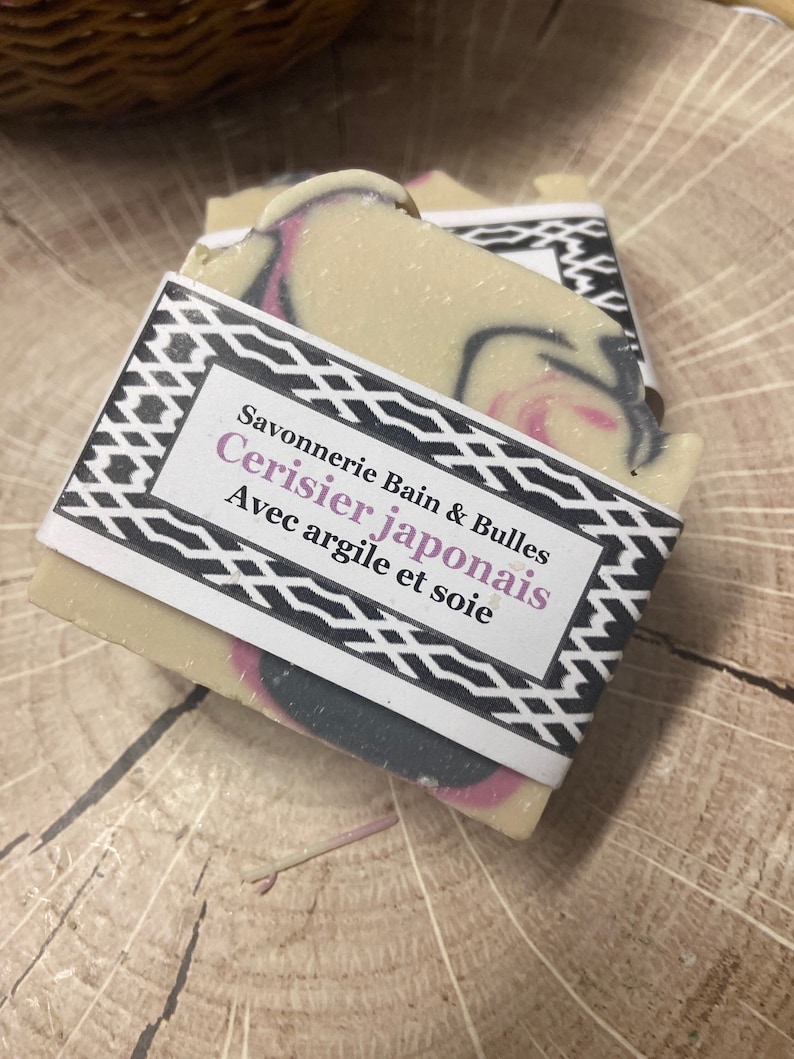 Puede incluir: Una pastilla de jab&oacute;n rectangular con una base de color crema que presenta remolinos de color rosa y gris oscuro. Una etiqueta de papel blanco envuelve el centro, impresa con el texto Savonnerie Bain & Bulles Cerisier japonais Avec argile et soie.