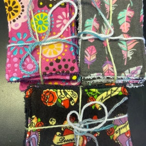 Op de afbeelding: Drie sets van stoffen vierkanten, elk gebonden met touw. De eerste set is roze met kleurrijke bloemenpatronen. De tweede set is grijs met witte en roze veren. De derde set is zwart met een tattoo-stijl ontwerp met harten, rozen en de woorden "Queen" en "Malefactor".