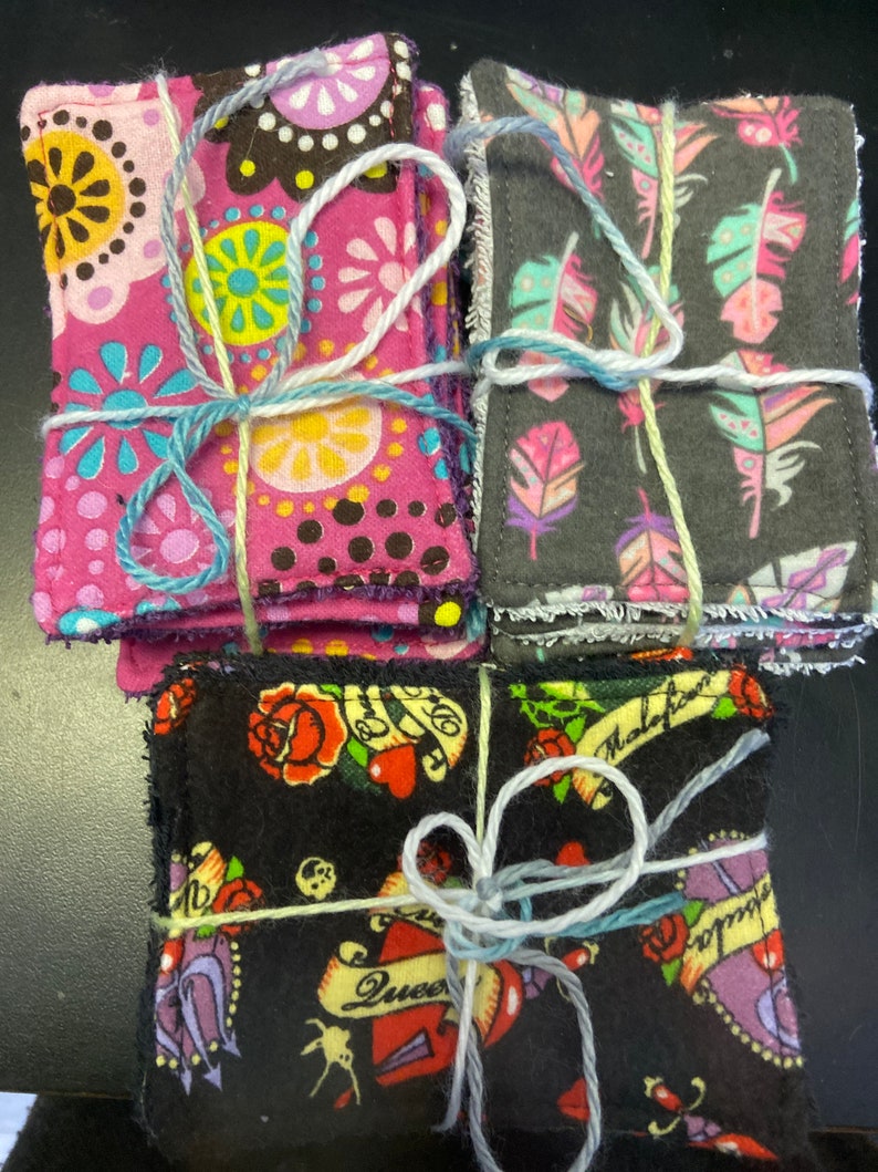 Op de afbeelding: Drie sets van stoffen vierkanten, elk gebonden met touw. De eerste set is roze met kleurrijke bloemenpatronen. De tweede set is grijs met witte en roze veren. De derde set is zwart met een tattoo-stijl ontwerp met harten, rozen en de woorden "Queen" en "Malefactor".