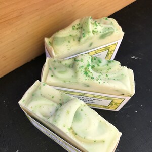 May include: Three bars of handmade soap with a light green and white swirl design, speckled with green granules. The soaps are in small, white paper trays with yellow and green labels. The background is a wooden surface and a black surface.