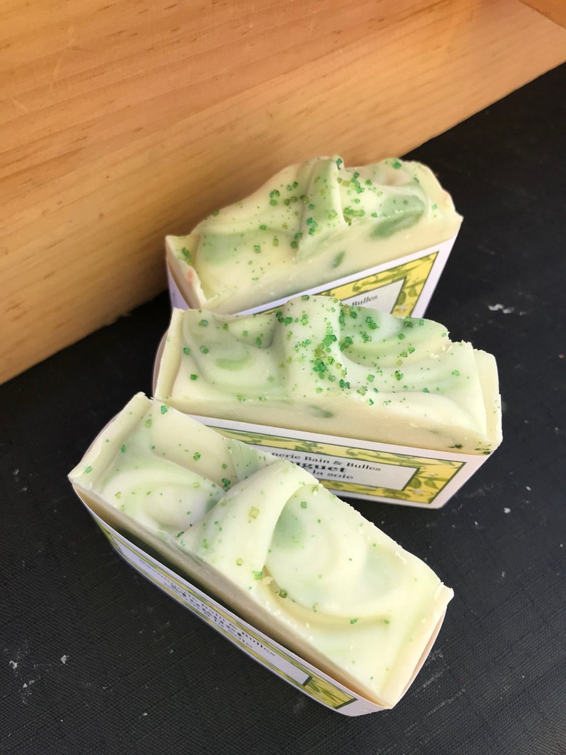 May include: Three bars of handmade soap with a light green and white swirl design, speckled with green granules. The soaps are in small, white paper trays with yellow and green labels. The background is a wooden surface and a black surface.