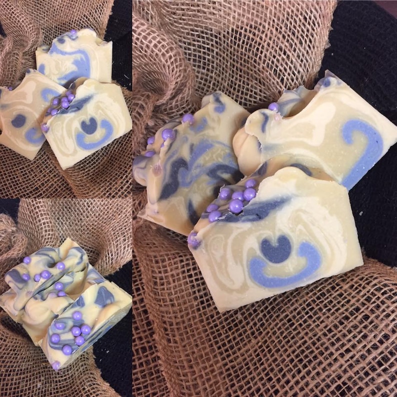 May include: Handmade soap bars with a creamy, marbled design. The soaps have blue and grey swirls, decorated with small purple beads. The bars are displayed on a burlap cloth, highlighting their artisan nature.