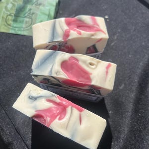 Puede incluir: Tres barras rectangulares de jab&oacute;n artesanal con un patr&oacute;n jaspeado de remolinos en color crema, gris carb&oacute;n y rosa vibrante. Las barras de jab&oacute;n tienen una textura suave y s&oacute;lida, y est&aacute;n dispuestas en una pila escalonada con una mezcla de colores uniforme.